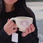 Person holding a ceramic cup outdoors with buildings and trees in the background